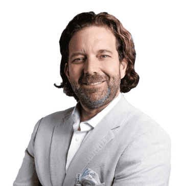 A professional businessman with shoulder-length brown hair and a beard, dressed in a light gray suit, smiling confidently against a plain background.